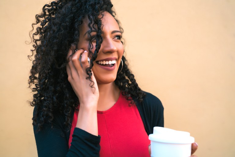 portrait of young afro woman talking on the phone while holding cup of coffee against yellow background communication co